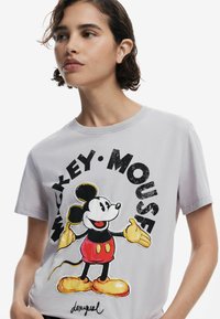T-shirt en coton gris avec un imprimé graphique de Mickey Mouse portant un short rouge et des chaussures jaunes, entouré de texte noir en gras.