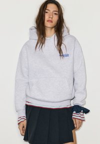 Grijze hoodie met voorzak, voorzien van een blauw logo; gecombineerd met een geplooide donkere rok; gestreepte manchetten met blauwe en rode accenten.