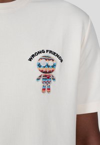 Krémové tričko s vyšitým motivem postavy, které má vzor modrých, červených a černých akcentů a text "WRONG FRIENDS" nad ním.