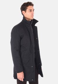 Cappotto lungo nero con colletto alto, chiusura con zip e bottoni a pressione. Include tasche laterali e uno strato interno trapuntato per l'isolamento.