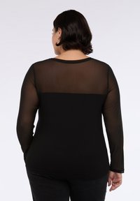 Donna vista da dietro che indossa un top nero a maniche lunghe con schiena superiore e maniche in rete trasparente, abbinato a pantaloni neri.