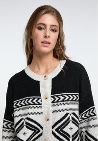 Schwarz-weißer gemusterter Cardigan mit rundem Halsausschnitt, großen beigen Knöpfen und strukturiertem Strick. Enthält geometrische Designs über das gesamte Teil.