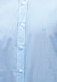 Κουστούμι με κάθετες ρίγες σε γαλάζιο και άσπρο, με λευκά κουμπιά και μικρό κεντημένο λογότυπο "NK" στο στήθος.