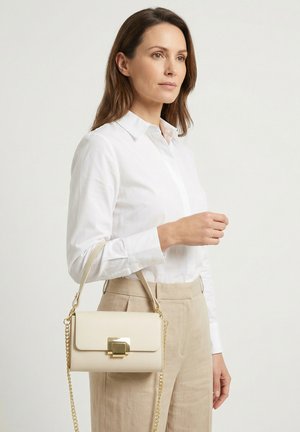 Vrouw in een wit overhemd met knopen en beige broek, met een kleine beige handtas met een gouden sluiting en kettingband.