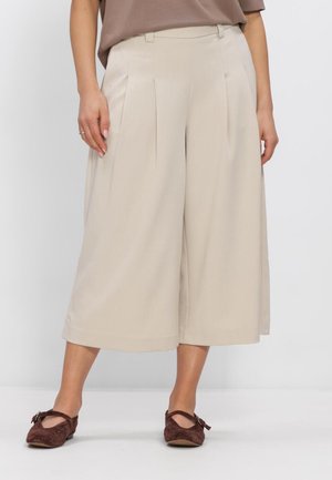 Femme portant un pantalon beige à jambes larges et courts et des ballerines en daim marron, debout devant un fond clair uni.