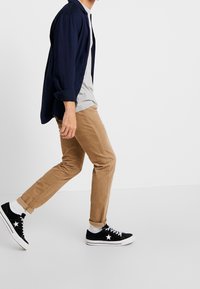 Námořnická košile s dlouhým rukávem přes šedé tričko, pískové slim-fit kalhoty a černé tenisky s bílou podrážkou a hvězdným detailem. Bílé ponožky jsou viditelné.