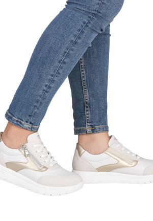 Baskets blanches et beige avec un accent métallique doré et un détail de fermeture éclair. Présentent une tige texturée et une semelle blanche. Portées avec un jean bleu.