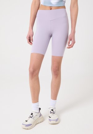 Mujer con shorts ciclistas de cintura alta color púrpura claro, calcetines blancos y zapatillas blancas con detalles en púrpura, de pie contra un fondo blanco.