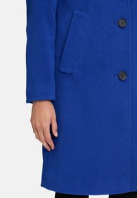 Ręka obok przedniej kieszeni, a guziki na rękawie niebieskiego wełnianego płaszcza widoczne na białym tle.