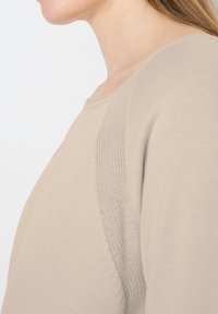 Gros plan sur le cou et l'épaule d'une personne portant un pull en maille beige texturé avec un col rond, sur fond blanc.