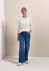 Hellgrauer Strickpullover mit strukturierter Oberfläche, kombiniert mit blauen hochtaillierten Schlagjeans und weißen Sneakers, vor einem neutralen Hintergrund.