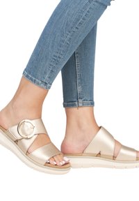 Gold metallic sandals with cross straps, circular buckle detail, textured finish, and a thick cream sole. Worn with blue denim jeans.