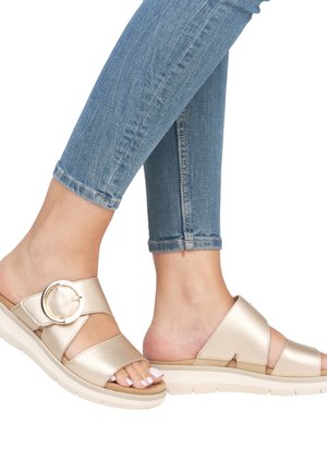 Sandales métalliques dorées avec des brides croisées, détail de boucle circulaire, finition texturée et épaisse semelle crème. Portées avec un jean en denim bleu.