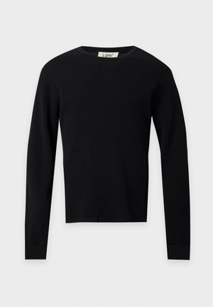 Homme debout, les mains dans les poches, portant une chemise noire à manches longues, un short bleu marine, des chaussettes blanches et des baskets marron sur fond blanc.