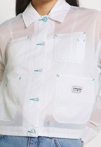 Giacca bianca trasparente con bottoni, cuciture turchesi, numerose tasche e una toppa con il logo Converse. Texture liscia, design leggero.