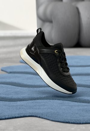 Zwarte geborstelde sneaker met witte zool die boven een blauw tapijt op een grijze vloer zweeft, met een wazige sculptuur op de achtergrond.