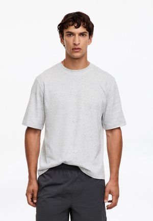 Jeune homme aux cheveux courts et bouclés, portant un t-shirt gris clair à manches courtes et un short noir, debout devant un fond blanc uni.
