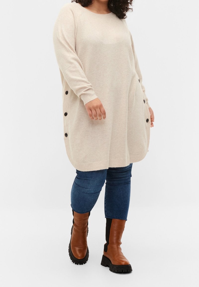 Personne portant un long pull beige avec des boutons noirs sur les côtés, un jean bleu et des bottes marron à semelle épaisse, marchant devant un fond uni.