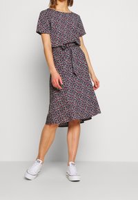 Blommig klänning med korta ärmar, marinblå botten med röda och vita blommor, insvängd midja med knytning, svängig kjol i kombination med vita sneakers.