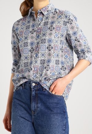 Persona que lleva una camisa de manga larga con botones y un diseño de azulejos en azul y rosa, y vaqueros azules de tiro alto con una mano en el bolsillo.