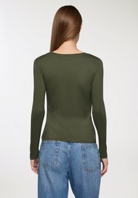 Długi rękaw w kolorze oliwkowym, wykonany z gładkiego materiału. Posiada okrągły dekolt oraz dopasowany krój, zestawiony z niebieskimi dżinsami o wysokim stanie.
