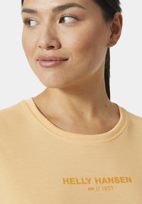 Košile s krátkým rukávem v broskvové barvě, s kulatým výstřihem a přiléhavým střihem, s logem "Helly Hansen" v kontrastní žluté barvě.