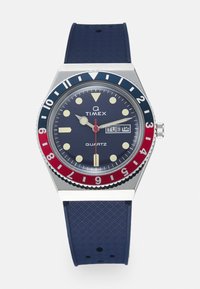 Timex quartz watch featuring a navy blue dial, silver stainless steel case, red and blue bezel, yellow hour markers, and rubber strap.