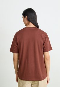 Bruine katoenen t-shirt met korte mouwen en een ronde hals, met een eenvoudige uitstraling en een ontspannen pasvorm. De achterkant toont een gladde textuur.