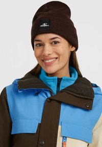 Hnědá pletená čepice s černou logovanou nášivkou; vrstvená bunda obsahuje modré, hnědé a béžové panely; viditelné zipové kapsy a detaily kapuce.