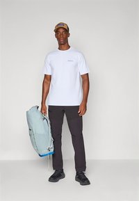 Hvit T-skjorte med korte ermer og blå logo, svarte bukser og en lys blå ryggsekk. Modellen har på seg en beige caps og svarte sko.