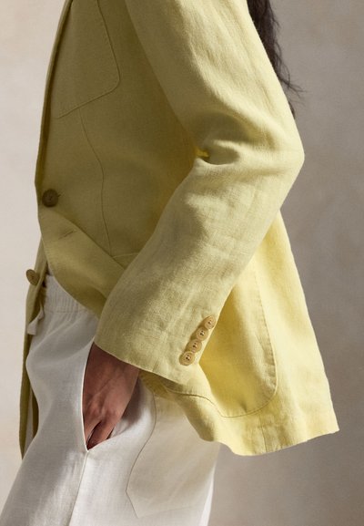 Personne portant un blazer en lin jaune avec des poignets boutonnés et un pantalon blanc, main posée dans la poche, vue de profil sur un fond neutre.
