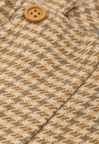 Tejido de punto en un patrón de pied-de-poule en beige, marrón y verde. Presenta un botón de madera, que muestra una superficie texturizada y una sensación suave.