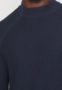 Zbliżenie na osobę w ciemnonawigacyjnym swetrze ze żłobionym splotem, z kołnierzem typu mock neck i rękawami raglanowymi.