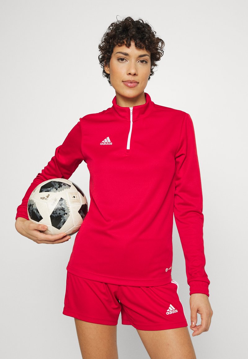 Conjunto atlético rojo que incluye una camiseta de manga larga con cremallera de cuarto y pantalones cortos a juego, ambos con detalles en blanco y el logo de Adidas. Balón de fútbol sostenido en la mano.