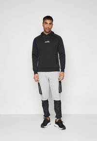 Black hoodie with a front logo, zip pockets, and grey sweatpants featuring black accents and two side pockets with green details. Black sneakers.