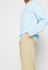 Sweatshirt bleu clair avec un col rond, des manches longues et un petit logo vert sur la poitrine ; associé à un pantalon beige.