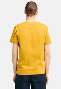 Hořčicově žluté tričko s krátkým rukávem a kulatým výstřihem, vyrobené z jemné a hladké látky. Zobrazena je zadní část, která ukazuje jednoduché designové prvky.