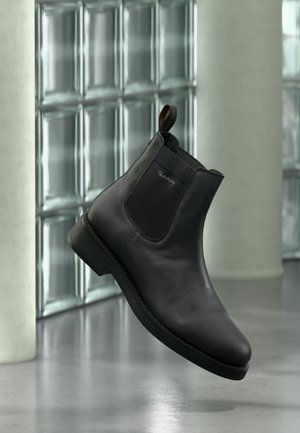 Bottine noire en cuir avec panneau latéral élastique et languette, présentée dans un cadre intérieur moderne avec un mur en blocs de verre en arrière-plan.