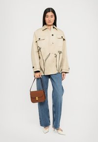 Giacca in cotone beige con stampa floreale nera, design oversize, tasche frontali, abbinata a jeans blu chiaro e una borsa marrone. Scarpe bianche a punta.