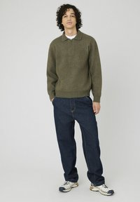 Pull zippé vert olive en texture tricotée, associé à un jean en denim bleu foncé ample et des baskets gris clair avec des accents noirs.