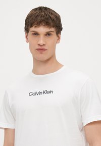 Bela bombažna majica s kratkimi rokavi in okroglim izrezom, z vezenim napisom "Calvin Klein" v črni barvi na prsnem delu.