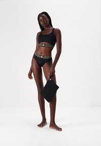 Μαύρο μπικίνι με χρυσό σχέδιο ελληνικού κλειδιού στις επάνω και κάτω ζώνες; μοντέλο κρατάει μια μαύρη τσάντα; απαλό ύφασμα, μινιμαλιστικός σχεδιασμός.