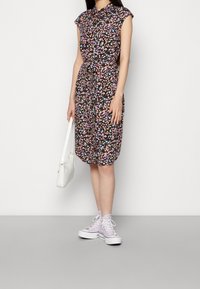 Robe midi fleurie avec fond noir, manches courtes et ceinture à la taille. Associée à un sac à main blanc et des baskets lilas.