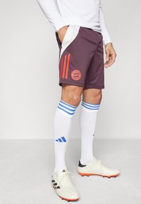 Shorts deportivos burdeos con detalles en naranja, que presentan un diseño de tres rayas, el logo del Bayern de Múnich y bolsillos laterales. Se llevan con calcetines blancos y botas de fútbol.