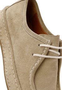 Zapato de ante beige con punta redonda, costuras visibles y cordones de color claro. Presenta un interior suave y una superficie texturizada.