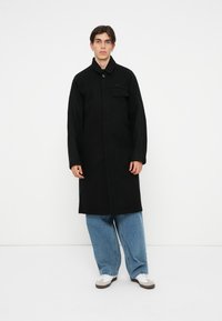 Cappotto nero realizzato in tessuto testurizzato, con chiusura a bottone, tasca sul petto e dettagli a coste alle maniche. Abbinato a jeans blu larghi e sneakers.