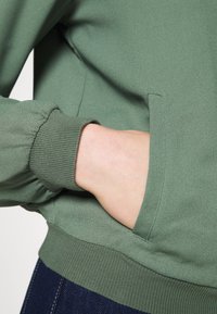 Zelená mikina s postranní kapsou, žebrovanými manžetami a hladkým povrchem, zobrazená na neutrálním pozadí.