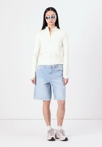 Hvit langermet zip-pullover med gule detaljer, lyseblå denimshorts og lyse joggesko med grå såle. Sølvfargede solbriller.