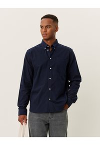Donkerblauw button-up shirt van katoen, met een borstzak, lange mouwen en witte knopen. Gepaard met grijze jeans en een tas.