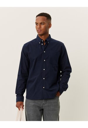 Navy blue button-up shirt made of cotton, featuring a chest pocket, long sleeves, and white buttons. Worn with gray jeans and a bag.